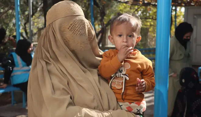 A woman holds her baby whose looking directly to camera whilst stood in front of some trees.