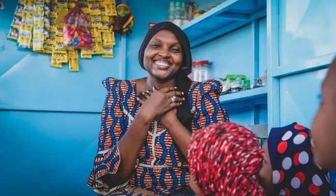 A smiling woman clasps her hands to her chest in front of her shop. 