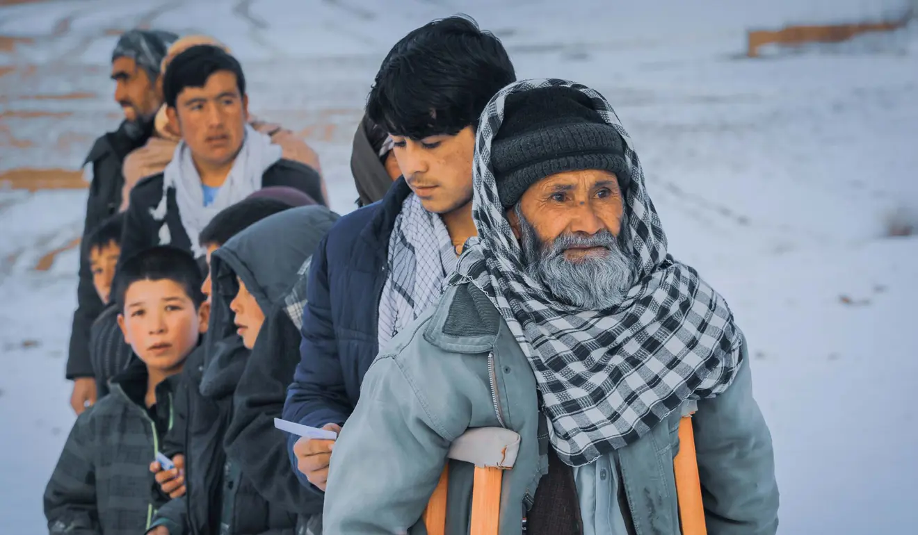 People in a queue, waiting for winter aid. The man at the front is elderly and using crutches. 
