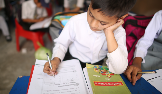 A child leans on their school desk whilst writing on paper at school. 