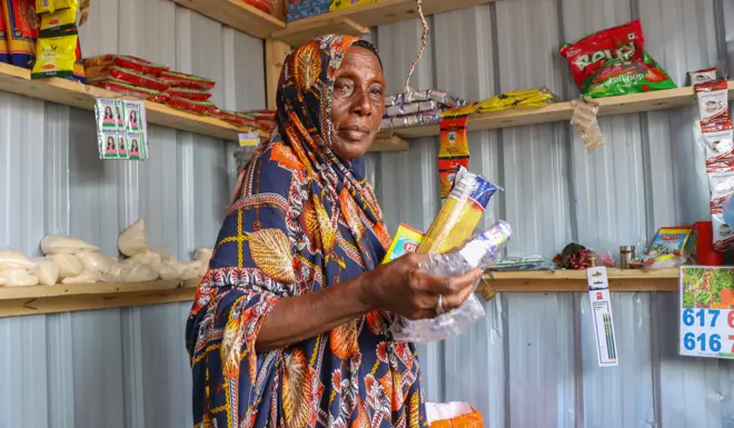 Amina stands in front of her shop where she is able to sell groceries. 